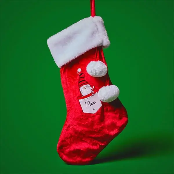 Personalised red Christmas stocking with a festive Santa design, white trim, and pom-pom details hanging against a green background.